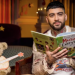 Former One Direction star Zayn Malik sits next to a teddy bear holding a toy guitar to read a