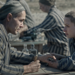 Lale (Jonah Hauer-King) tattoos Gita’s (Anna Próchniak) arm as they sit across each other at a table wearing striped gray prisoners’ uniforms in “The Tattooist of Auschwitz”