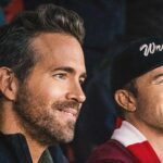 Ryan Reynolds and Rob McElhenney watching a game at the Wrexham stadium in Wales, draped in red and white club colours.