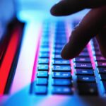 A zoomed-in image of a hand typing on a laptop. The laptop is bathed in red and blue light