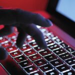 A hand typing at a computer in a dark room, lit up by the laptop