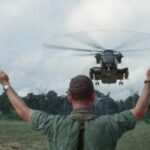 A man stands with his arms to the air as a helicopter approaches Jonestown