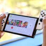 Nintendo Switch OLED held between two hands with one of the JoyCons being slid off