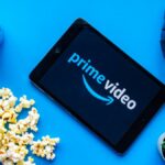 Prime Video logo appears on a tablet surrounded by a can of soda, spilled popcorn, headphones and a cactus