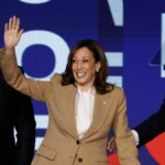 Kamala Harris, wearing a tan suit and flanked by Doug Emhoff and Joe Biden, waves to the crowd at the Democratic National Convention 2024