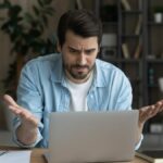 A man frowning and gesturing in a frustrated manner at his laptop