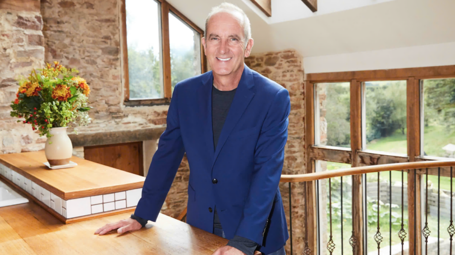 Kevin McCloud, wearing a royal blue jacket, smiles in an airy but rustic cottage kitchen as he presents &quot;Grand Designs: 25 Years and Counting&quot;