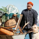 Banjo Beale, wearing red woolly hat and workmanlike denim jacket, carries a wheelbarrow filled with bric-a-brac alongside his faithful dog in BBC's Designing the Hebrides season 2