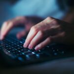 A close-up photograph of a person's hands typing on a backlit laptop keyboard