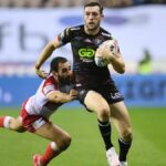 Jake Wardle, in all black kit, skips a challenge from Tyrone May, in red and white, in Wigan vs Hull KR.