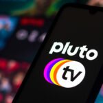 Pluto TV logo seen on a phone in front of a screen of blurry TV show images