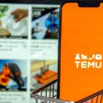 A phone with the Temu logo sits in a shopping cart in front of a computer screen with Temu items on it.