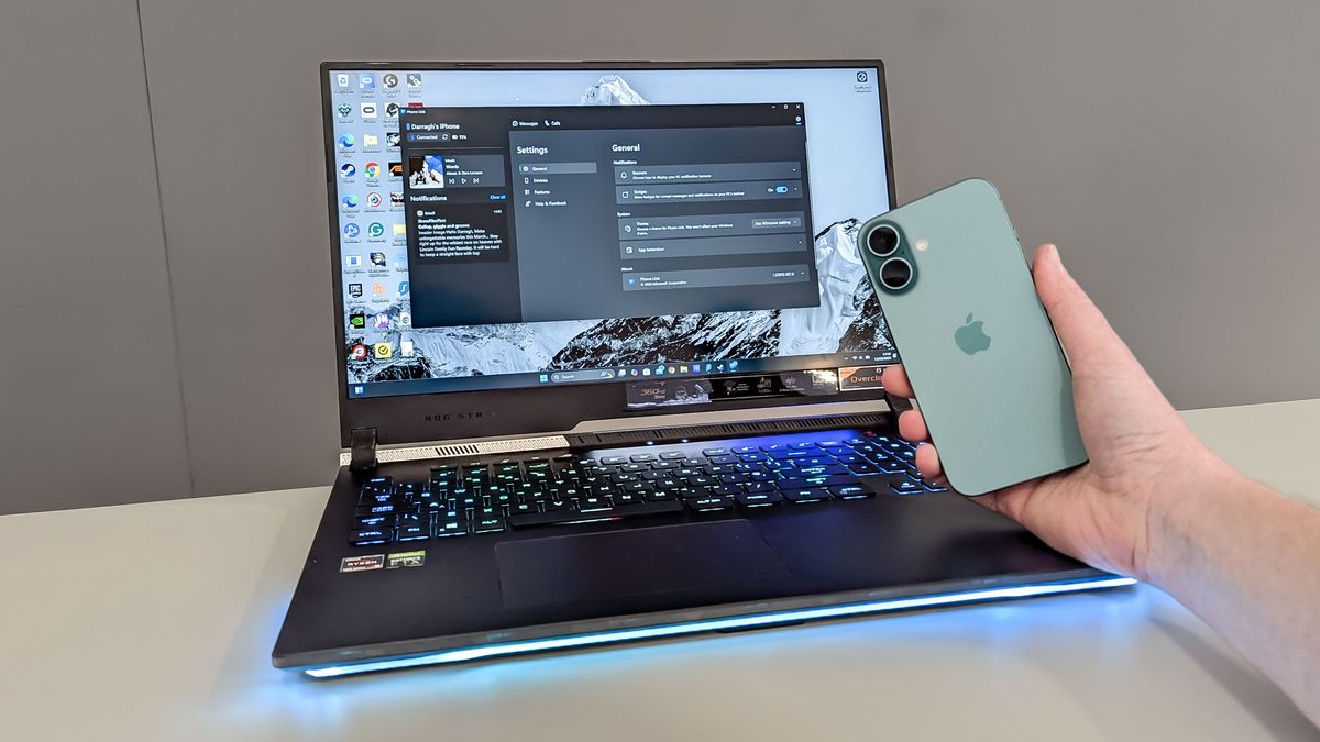 من آیفون 16 خود را با رایانه شخصی ویندوز هماهنگ کردم - و اتصال تلفن مایکروسافت نمی تواند با MacBook رقابت کند iPhone 16 in hand being held in front of Asus ROG Strix 17 on table showing Phone Link app